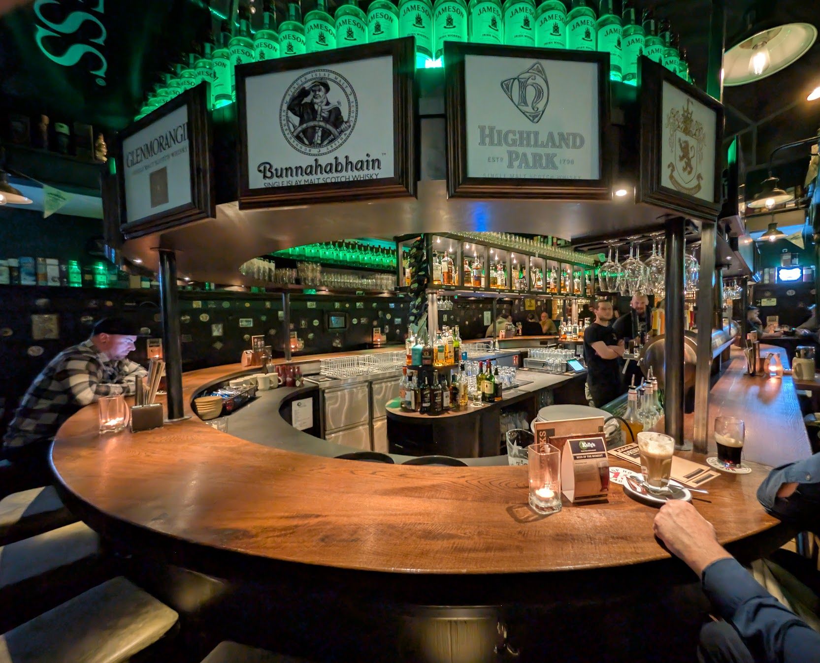 Zentrale, runde Holztheke des Kelly's Irish Pub in Ulm. Über der Bar sind Schilder von schottischen Whiskymarken (Glenmorangie, Highland Park) angebracht und leuchtende Jameson-Flaschen dekorieren die Decke.