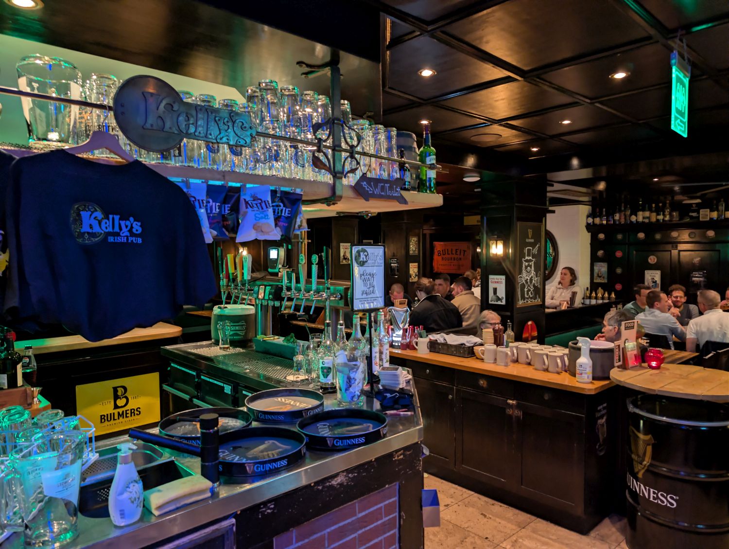 Das Erdgeschoss des Kellys Irish Pub in Ulm. Zu sehen ist die Bar mit Guinness-Tabletts, ein Stehtisch-Fass mit Guinness Logo und eine Gruppe Gäste im Hintergrund. An der Bar hängt ein T-Shirt mit dem Logo von Kellys Pub.