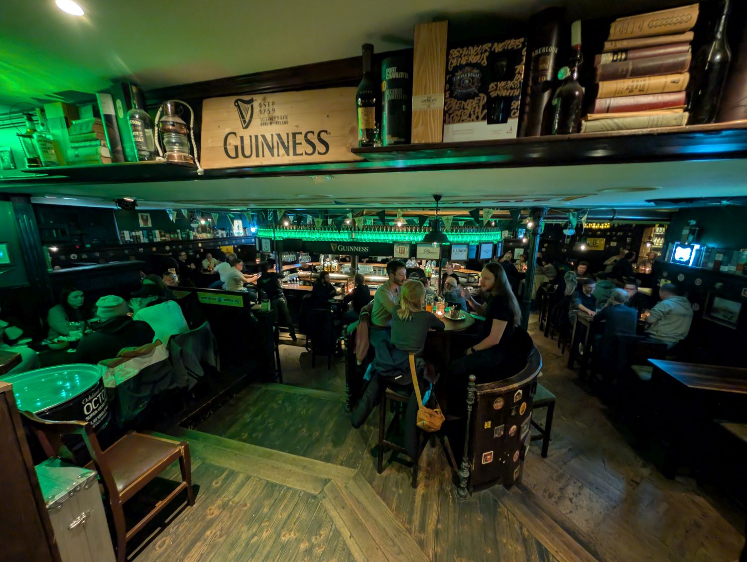 Innenansicht Kelly's Irish Pub in Ulm. Der große Raum wird durch dunkle Holzeinrichtung, eine Bar mit Guinness-Dekor und grüner Beleuchtung optisch gemütlicher und intimer gestaltet.